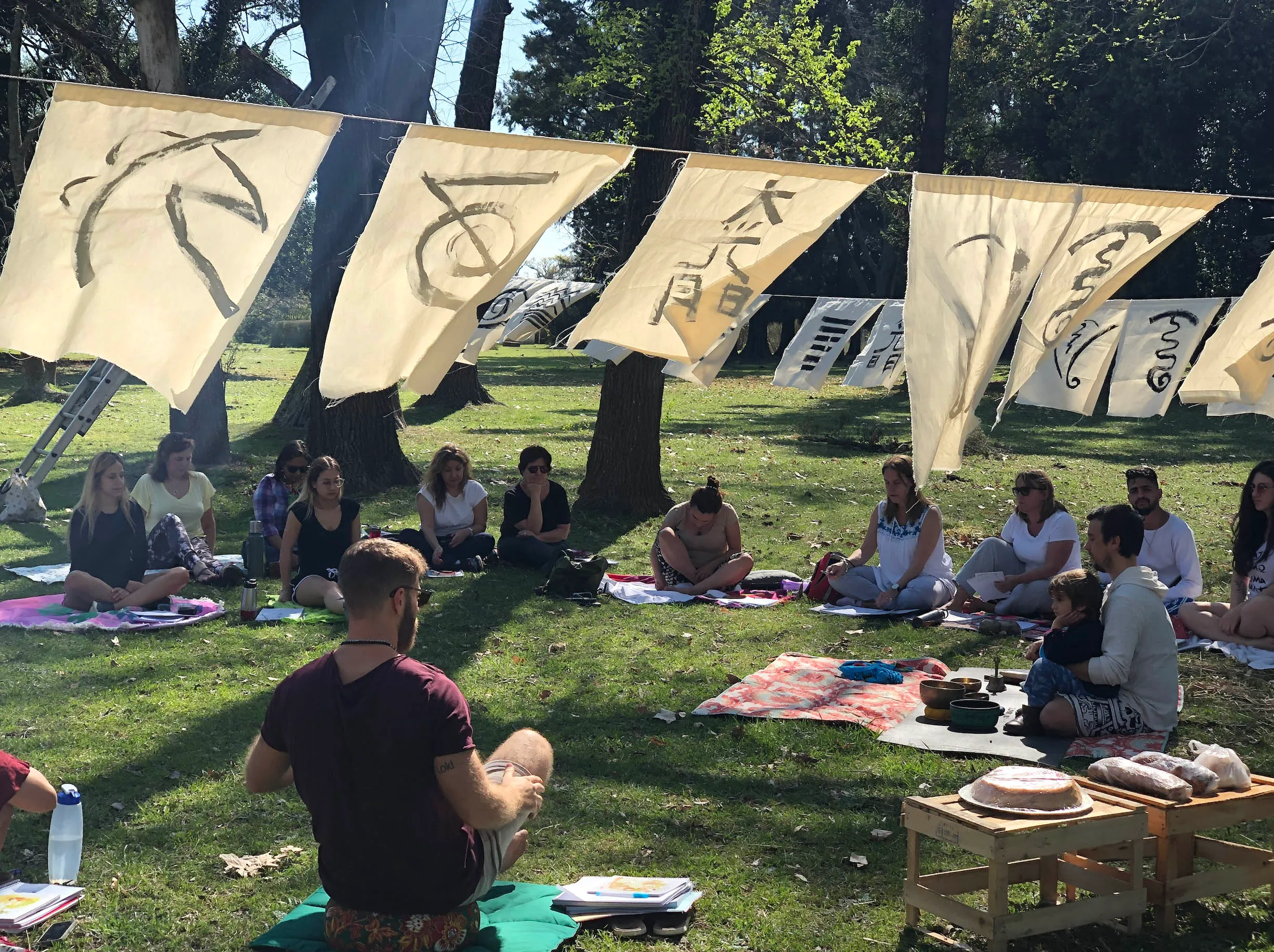 Mati de espaldas con tambor medicinal, kanji colgados con el sol detrás, círculo de practicantes meditando